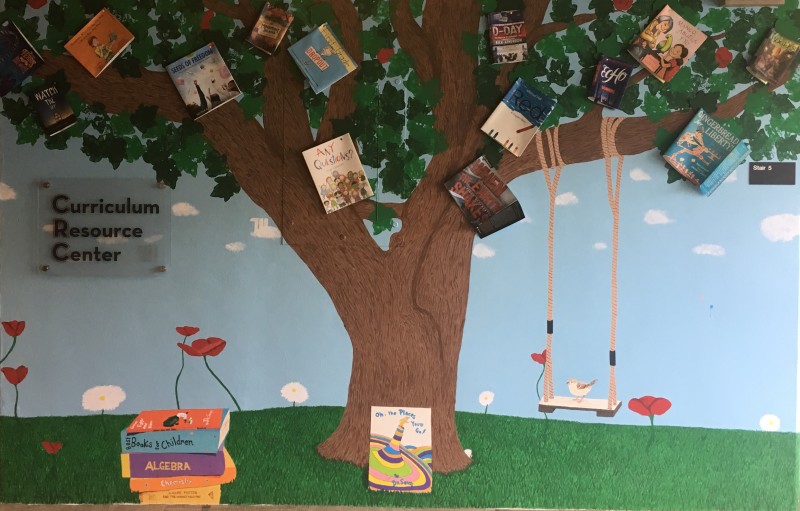 CRC Mural: depicts a tree in a field. There are book covers scattered among the leaves. There is a swing hanging from a branch, and a bird perched on the swing. A stack of books sits next to the tree, and a frog sits atop the books.